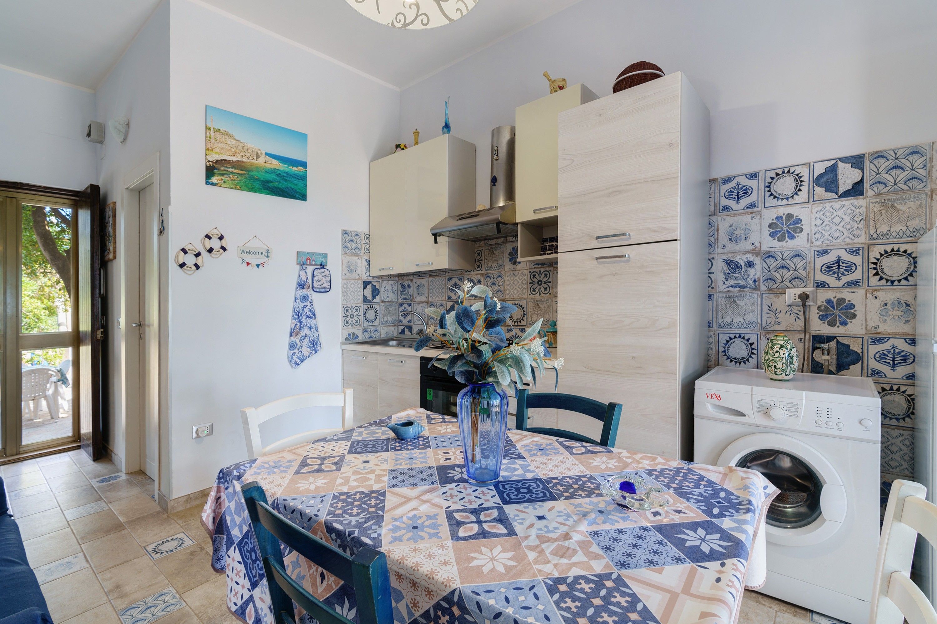 Une cuisine compacte avec un coin repas, comprenant un lave-linge, un pare-feu en carrelage et une vue sur l’extérieur grâce à une porte ouverte.