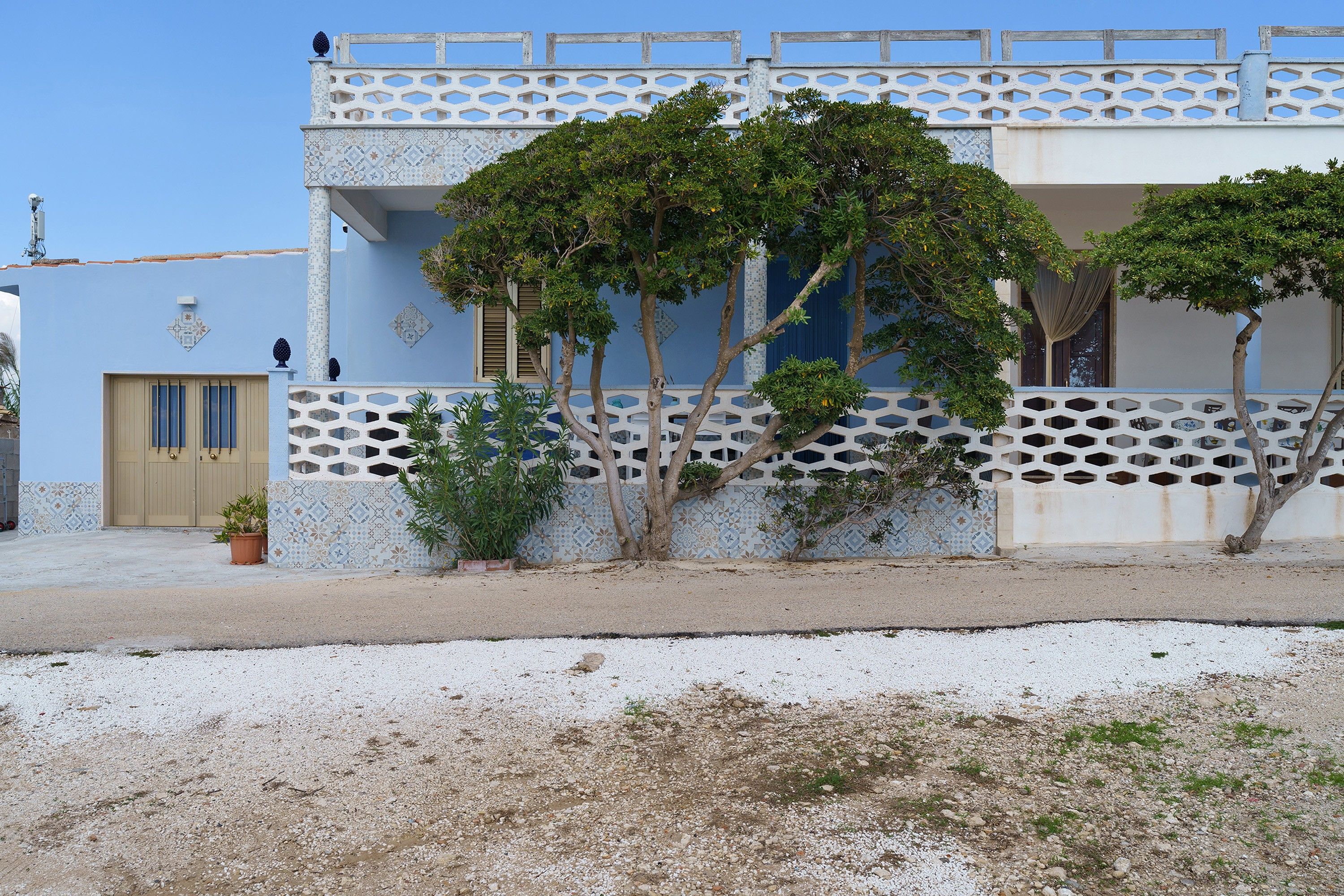 Un edificio con un muro exterior con diseño, una entrada protegida por una reja y un pequeño jardín con plantas en macetas.