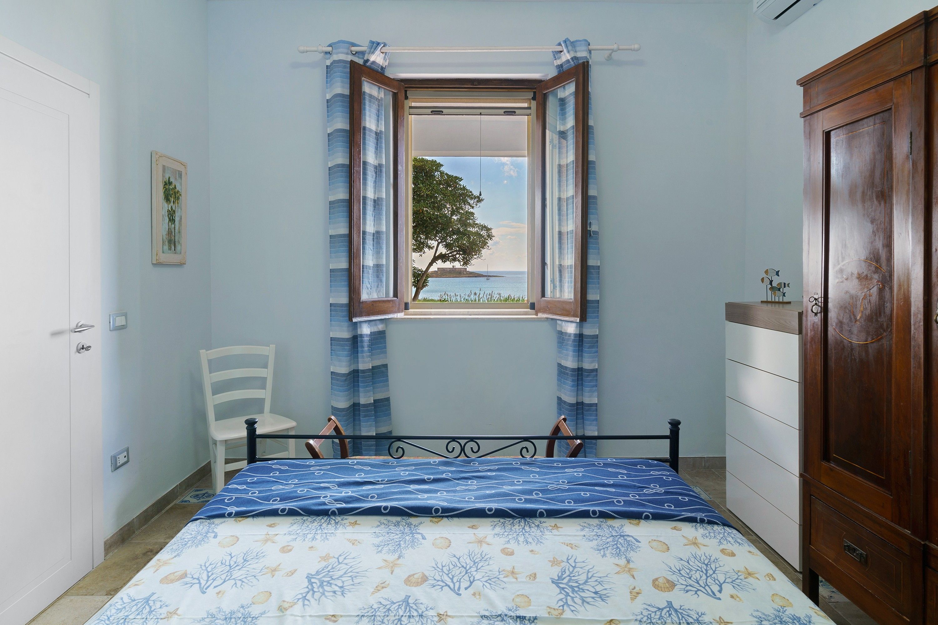 Ein Schlafzimmer mit Bett, Sessel, Kleiderschrank, einem Fenster mit Vorhängen und einem Bild an der Wand.