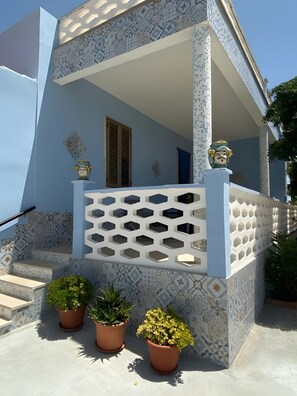 Un bâtiment à la façade bleue, doté d’un balcon blanc orné d’un motif géométrique en treillis et de plantes en pot à l’entrée.