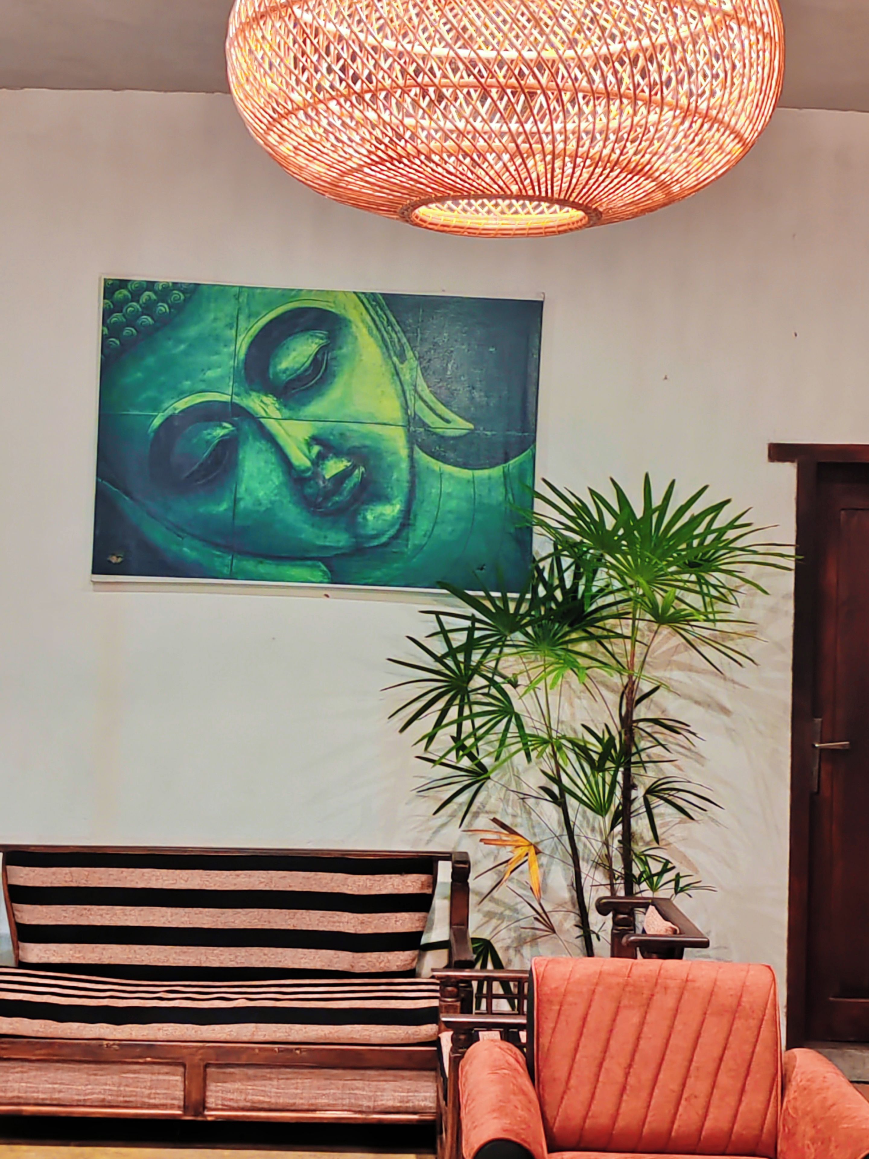 A room with a large Buddha painting, a wooden sofa, a striped chair, a potted plant, and a hanging light fixture.