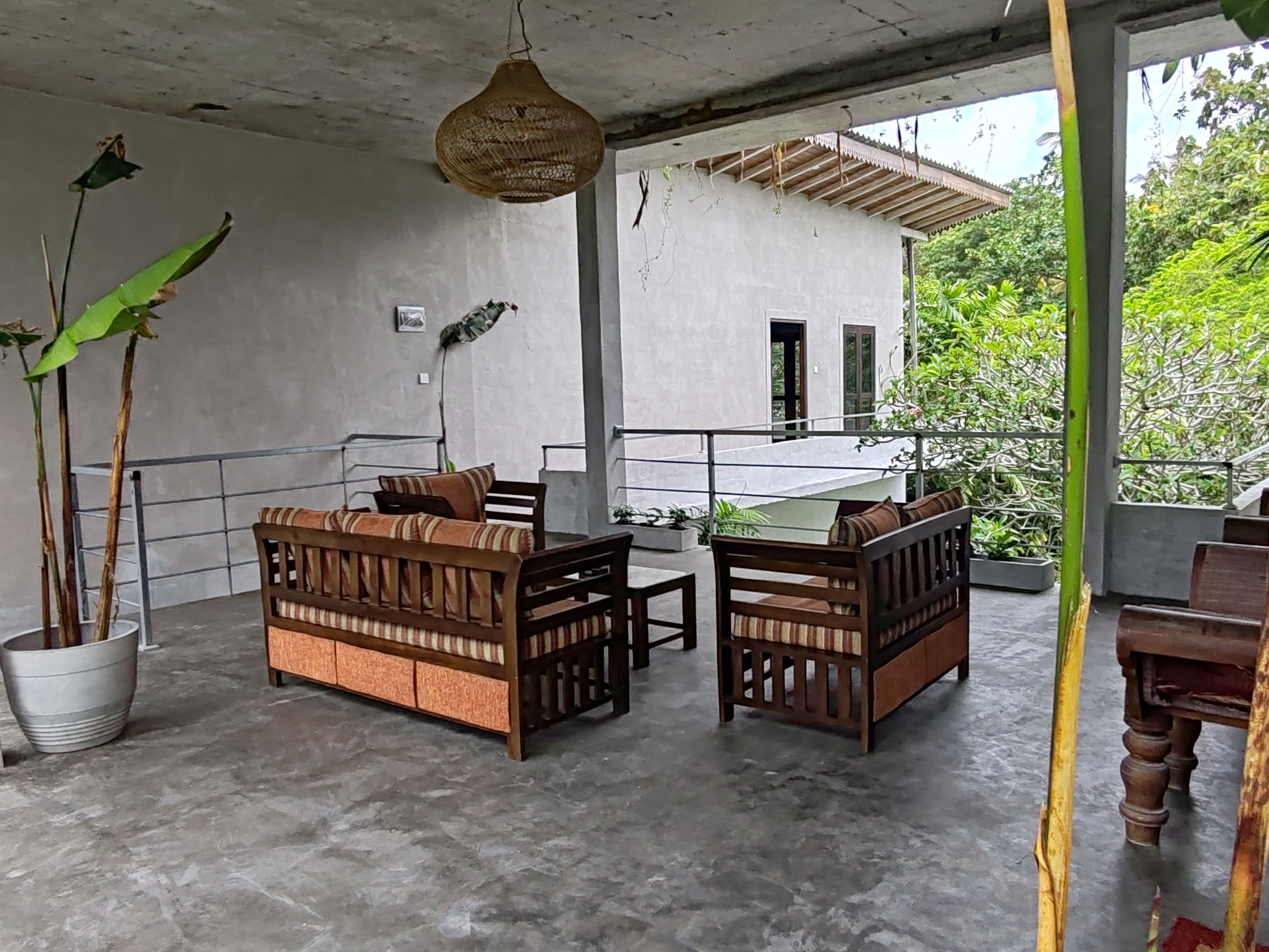 Un espace extérieur aménagé, abrité et doté de deux canapés en bois et d’une table, entouré de plantes en pot et agrémenté d’un luminaire suspendu.