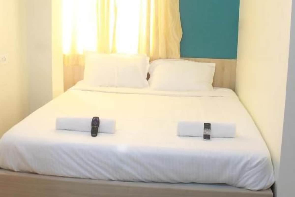 A neatly made bed with white linens and two pillows, an air conditioning unit mounted on the wall, and a window with curtains.