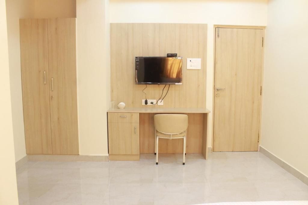 A room with a wooden wardrobe, a desk with a chair, and a mounted television.