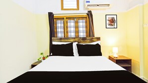 Hotellihuone, jossa on sänky, yöpöytä, lamppu, ikkuna verhoilla ja seinällä oleva kehystetty kuva.