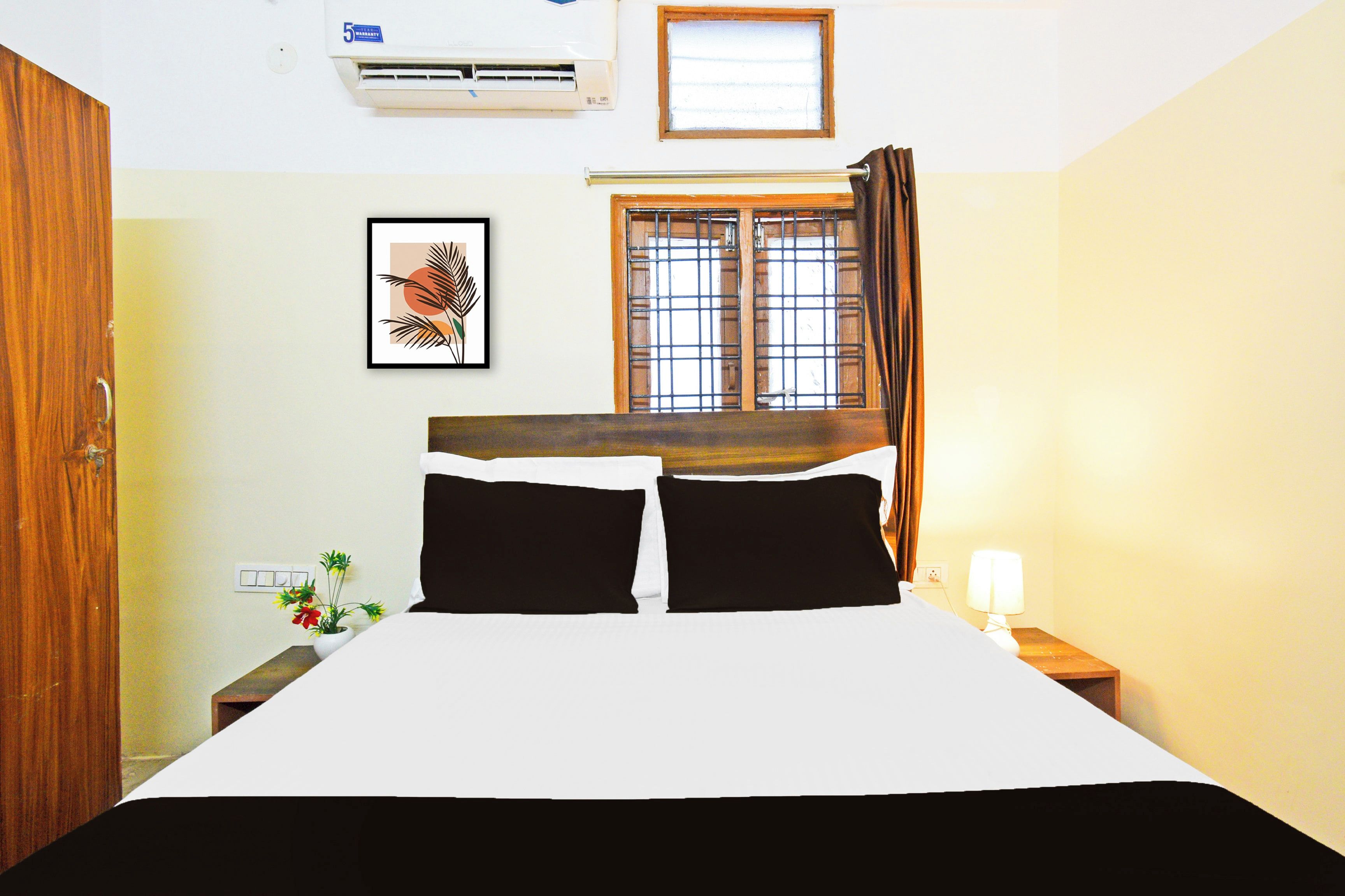 Una camera d'albergo con un letto, una finestra con tende, un condizionatore d'aria a parete, un quadro incorniciato e un comodino con una lampada.