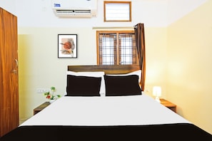 Quarto de hotel com cama, janela com cortinas, ar condicionado fixado na parede, quadro emoldurado e mesa de cabeceira com abajur.