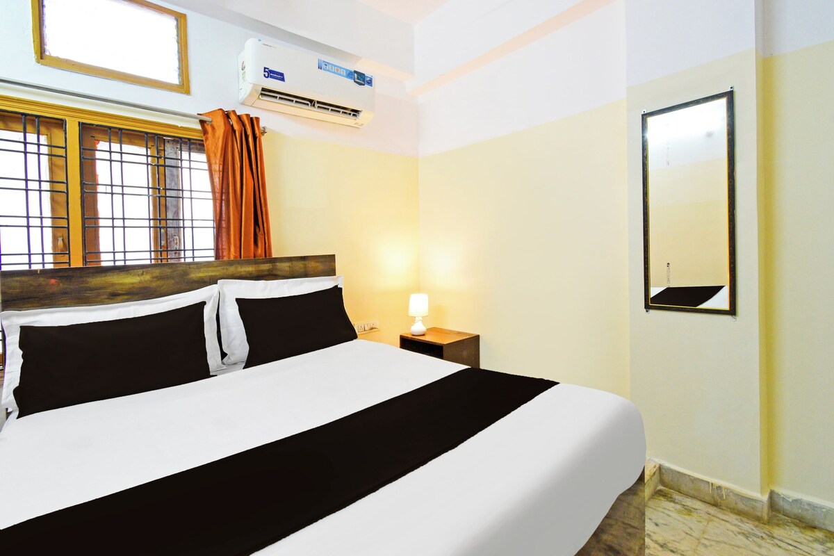 Une chambre d’hôtel comprenant un lit, une table de chevet avec une lampe, un appareil de climatisation, une fenêtre avec des rideaux et un miroir fixé au mur.