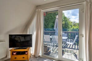Quarto com televisão sobre suporte de madeira, porta de vidro deslizante para um deck externo com cadeiras e guarda-sol, e um aviso de 'Não Fumar' na porta.