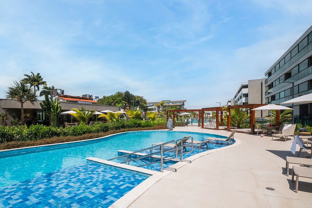 Piscina com borda azulejada, cercada por espreguiçadeiras e guarda-sóis, com edifícios ao fundo.