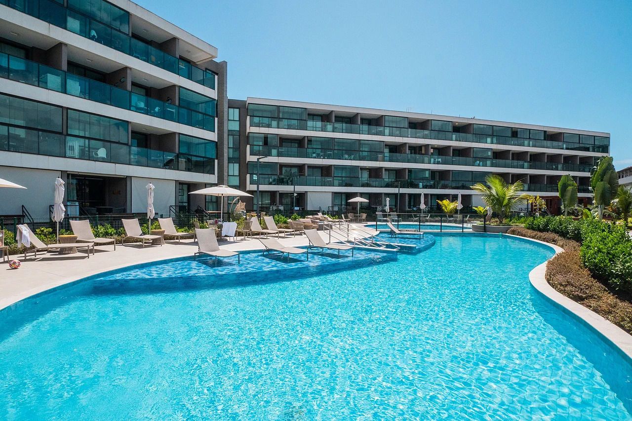 Una piscina con sillones y sombrillas, rodeada por un edificio moderno con varios balcones.