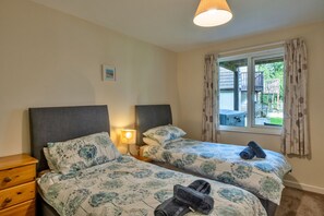 Une chambre avec deux lits, une table de chevet, une lampe, une fenêtre avec des rideaux et une petite commode en bois.