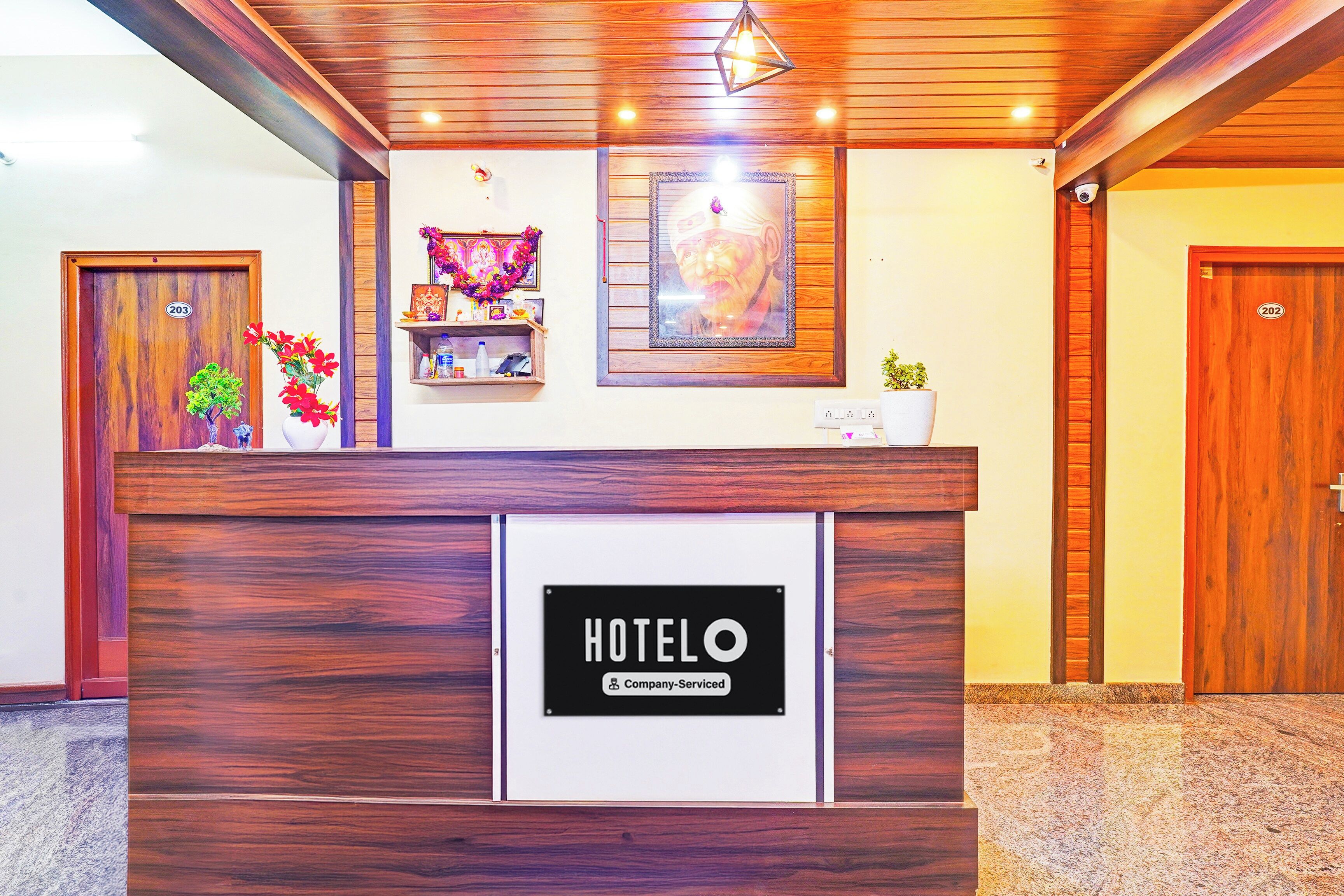 Un comptoir de réception d’hôtel avec un panneau indiquant qu’il est géré par l’entreprise. On y voit des portes en bois, une étagère fixée au mur avec divers objets, et un tableau encadré au-dessus du comptoir.