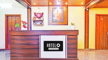 Un comptoir de réception d’hôtel avec un panneau indiquant qu’il est géré par l’entreprise. On y voit des portes en bois, une étagère fixée au mur avec divers objets, et un tableau encadré au-dessus du comptoir.