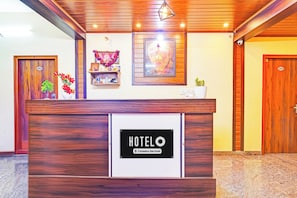 A hotel reception desk with a sign indicating it's company-serviced. There are wooden doors, a wall-mounted shelf with items, and a framed picture above the desk. - Hotel O Srigandhakavalu Formerly (Bengaluru)