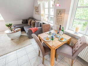 Ein gemütlicher Essbereich mit einem Holztisch, gedeckt für zwei Personen, einem grauen Sofa und einem kleinen Beistelltisch mit einer Vase und einem Weinglas.