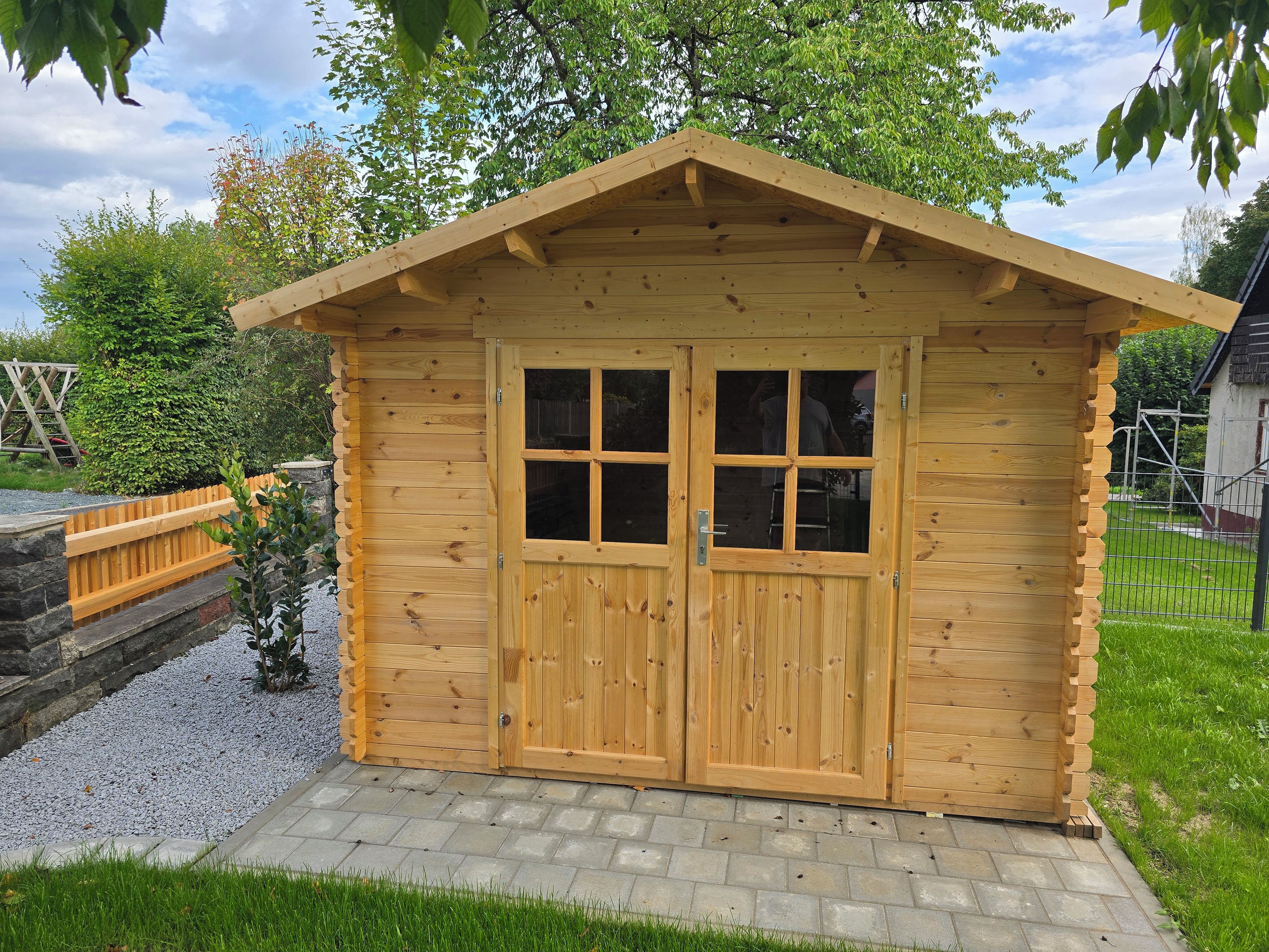 Eine Holzlagerhütte mit Satteldach und doppeltürigem Eingang, umgeben von einem Kiesweg und Grünpflanzen.