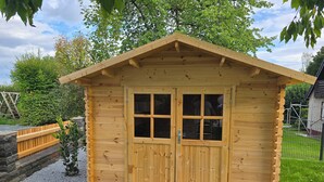 Ett trähus med ett sadeltak och en dubbeldörr, omgivet av en grusgång och grönska.