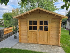 Eine Holzlagerhütte mit Satteldach und doppeltürigem Eingang, umgeben von einem Kiesweg und Grünpflanzen.