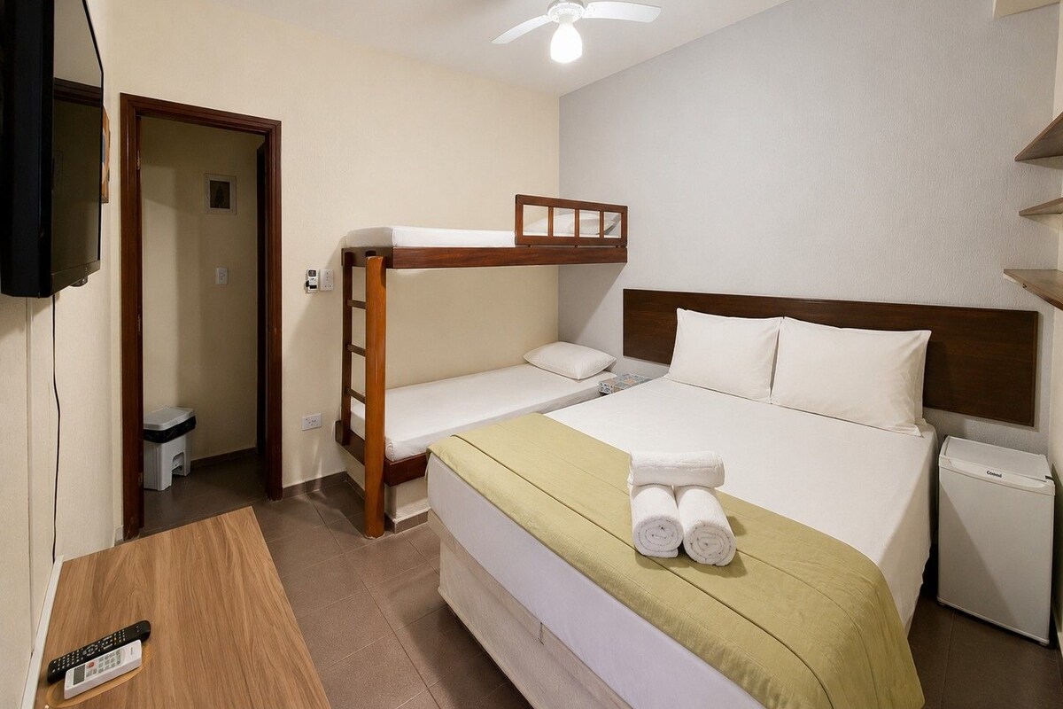 Une chambre d’hôtel équipée d’un lit superposé, d’un lit simple, d’une table de chevet, d’une télévision, d’un ventilateur de plafond et d’un petit radiateur de chevet.