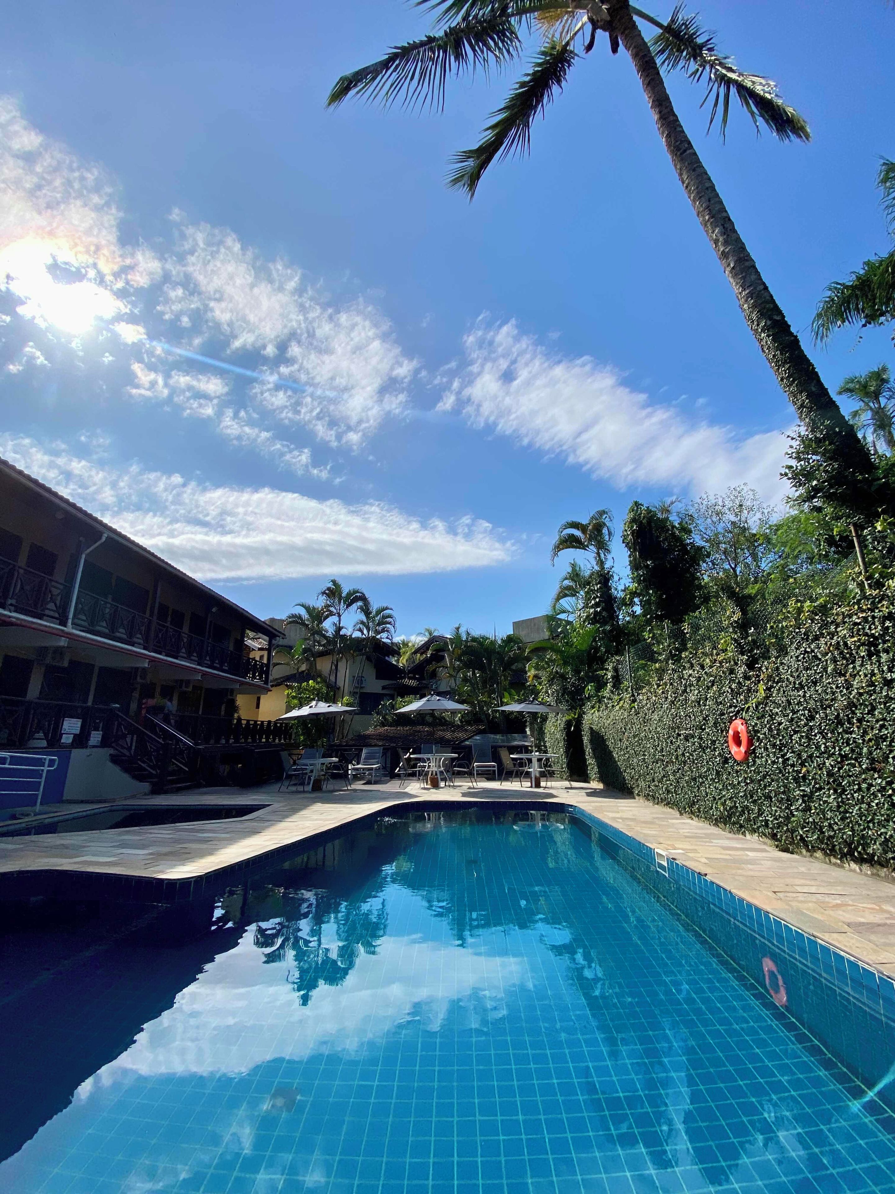 Piscina com piso ladrilhado, rodeada por espreguiçadeiras e guarda-sóis, com um edifício com varanda ao fundo.