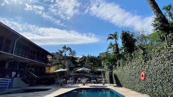 Piscina com piso ladrilhado, rodeada por espreguiçadeiras e guarda-sóis, com um edifício com varanda ao fundo.