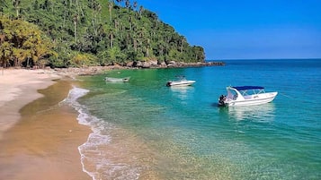 Une plage à l’eau turquoise et limpide, une rive sablonneuse et, en arrière-plan, une colline boisée.