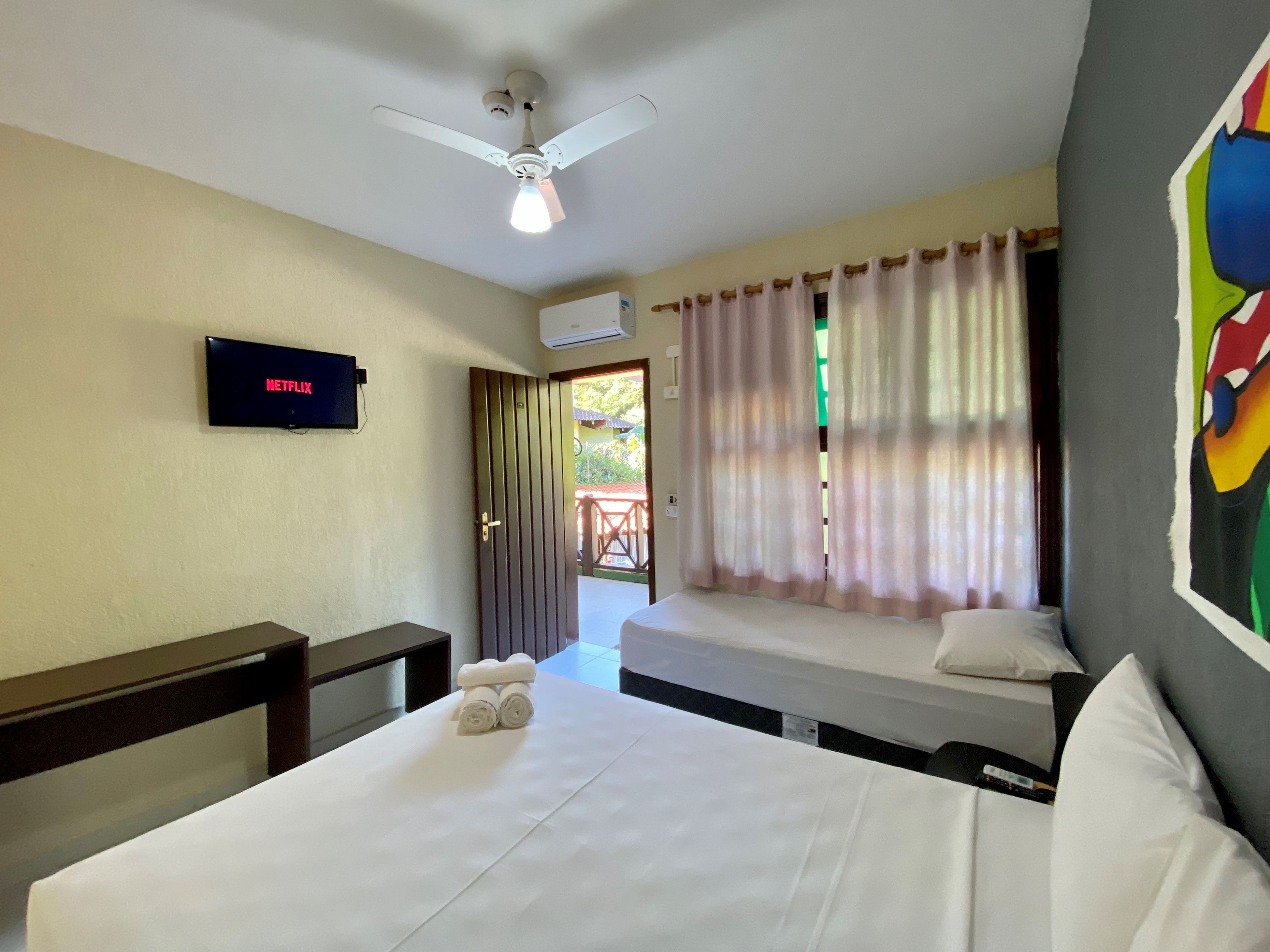 A hotel room with a bed, a TV mounted on the wall, a ceiling fan, and a window with curtains.