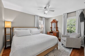 Une chambre d’hôtel avec un lit, une commode, une chaise, un ventilateur de plafond et une fenêtre avec des rideaux.