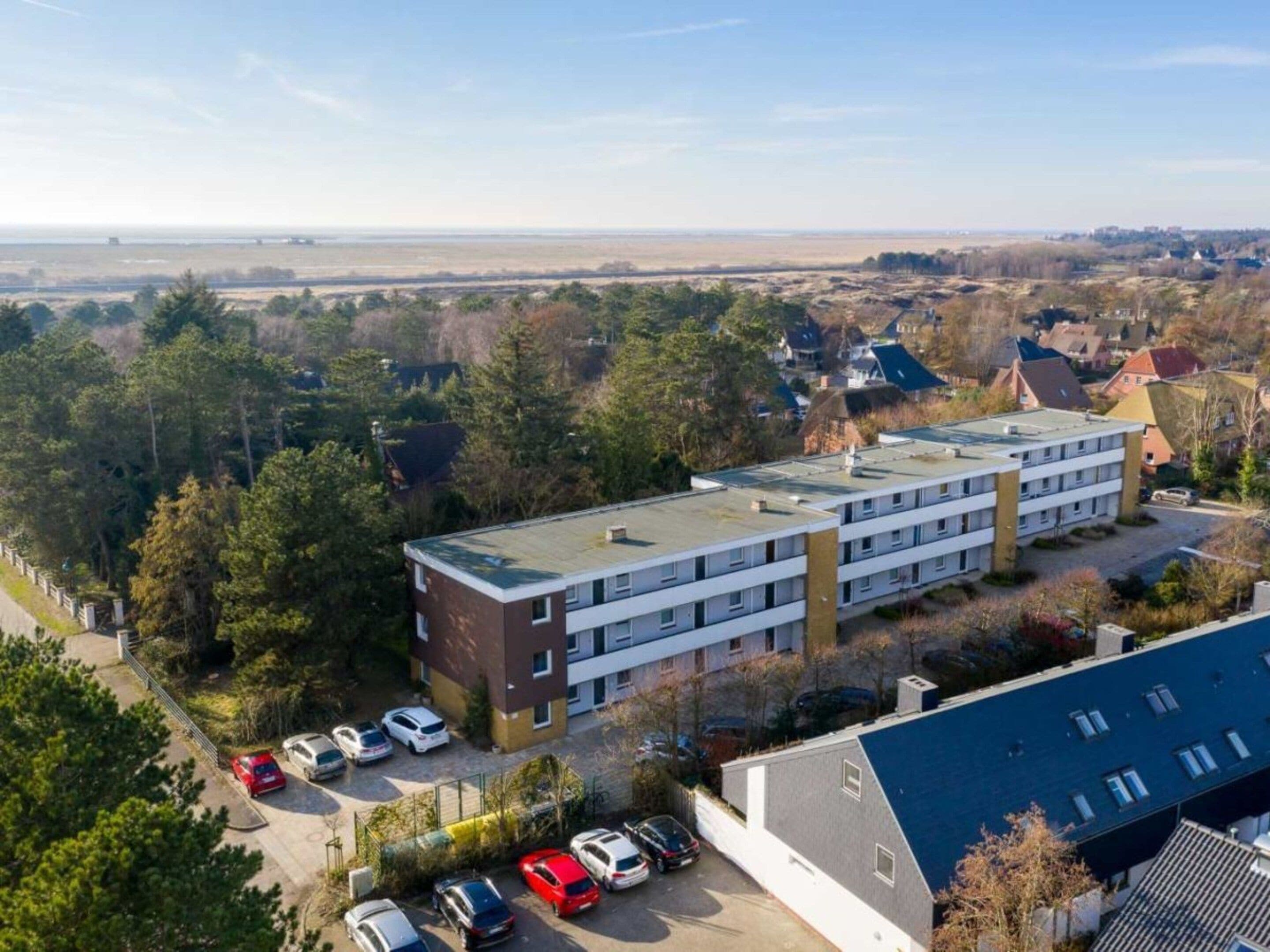 A multi-story building with a flat roof, surrounded by trees and a parking lot with several cars.