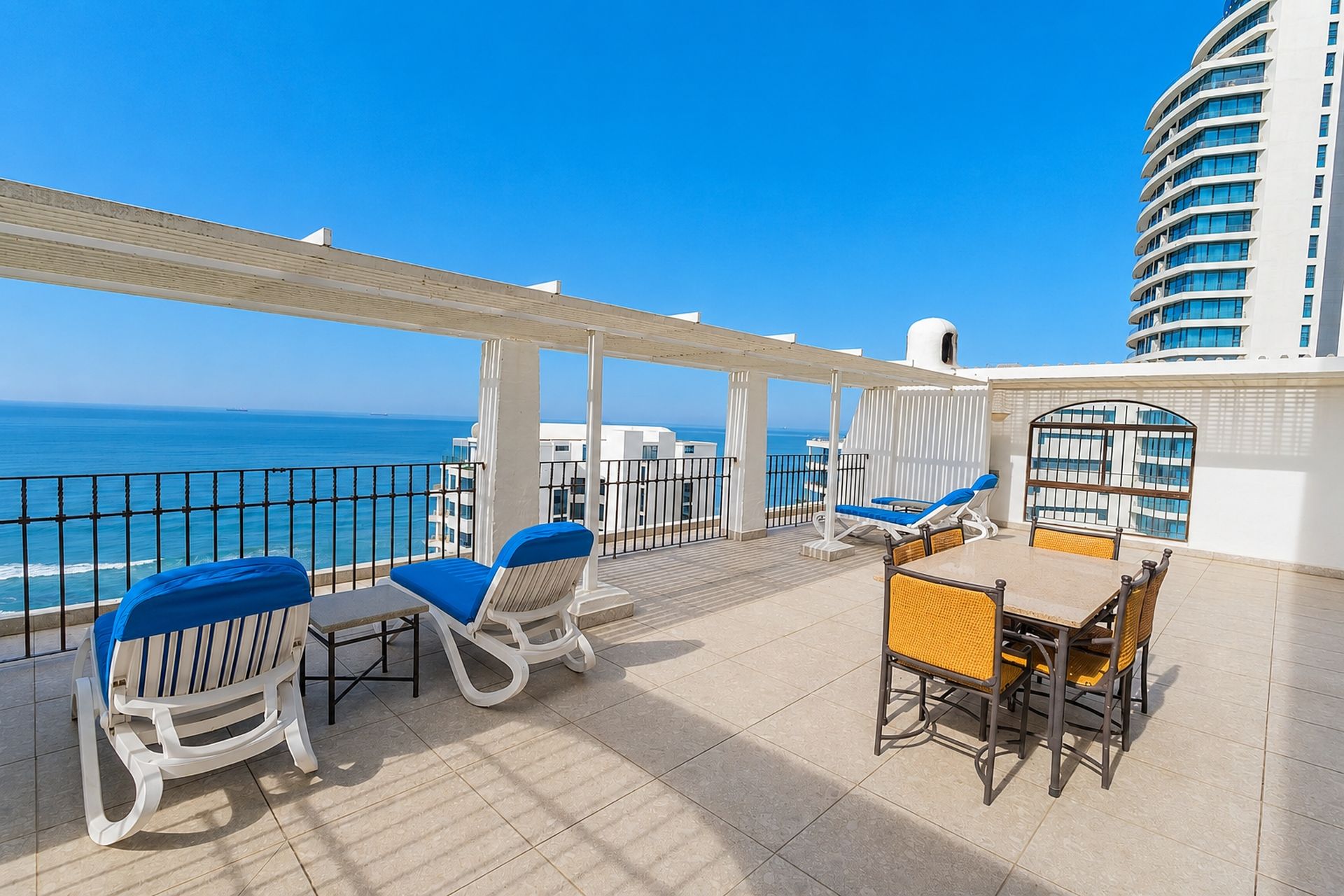 A sunlit outdoor seating area with a view of the sea, featuring white and blue lounge chairs and a table.