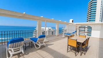 A sunlit outdoor seating area with a view of the sea, featuring white and blue lounge chairs and a table.