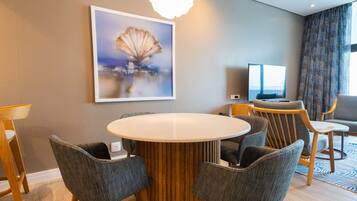 A hotel room with a round dining table, six grey upholstered chairs, a television, and a painting of a tree on the wall.