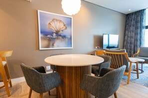Habitación de hotel con una mesa de comedor redonda, seis sillas grises tapizadas, un televisor y un cuadro de un árbol en la pared.