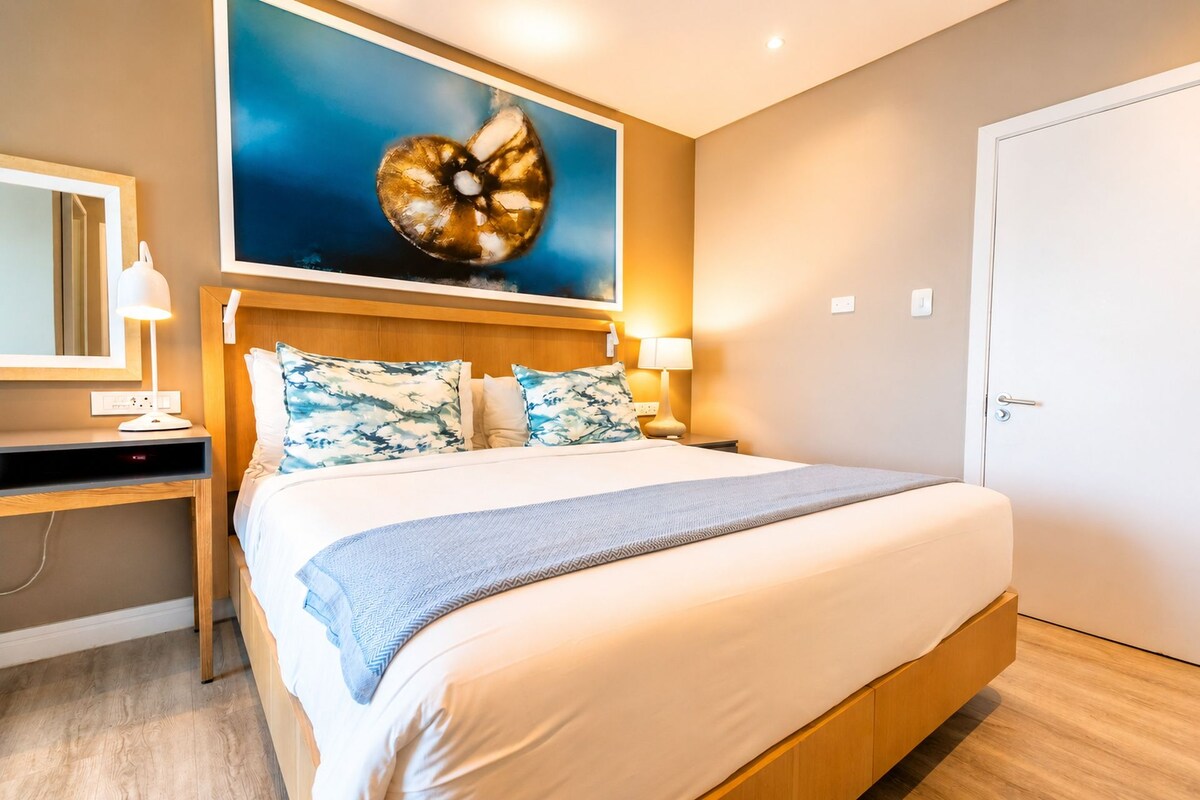 A hotel room with a bed, bedside table, lamp, and a painting on the wall.