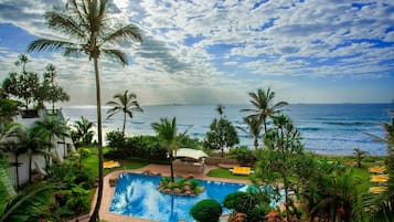 A pool area with a view of the ocean, surrounded by palm trees and a white building.