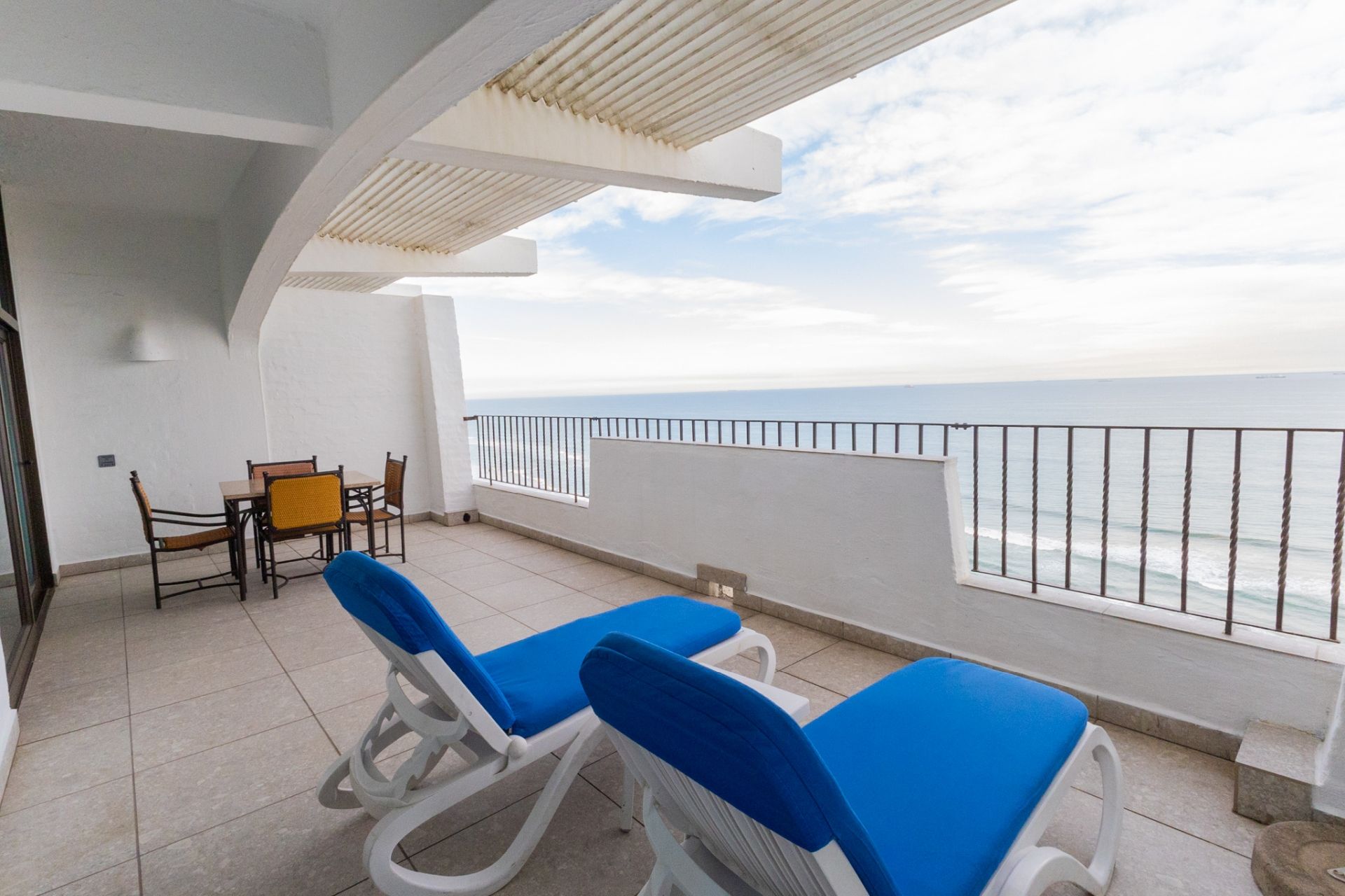 A balcony with two blue lounge chairs, a small table with chairs, and a view of the sea.