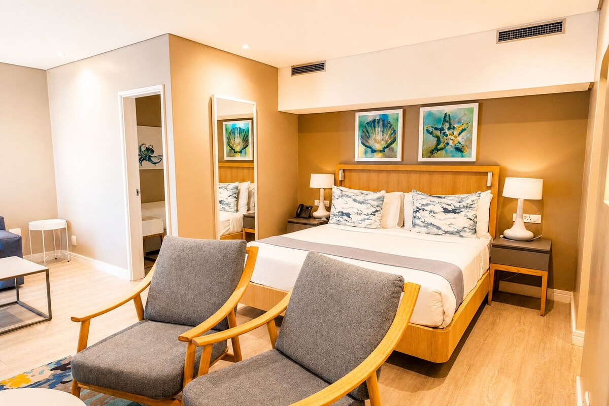 A hotel room with a bed, two armchairs, a bedside table with a lamp, and artwork on the wall.