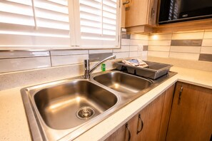 Una cocina con un lavamanos de acero inoxidable doble, un microondas encima y una ventana con persianas.