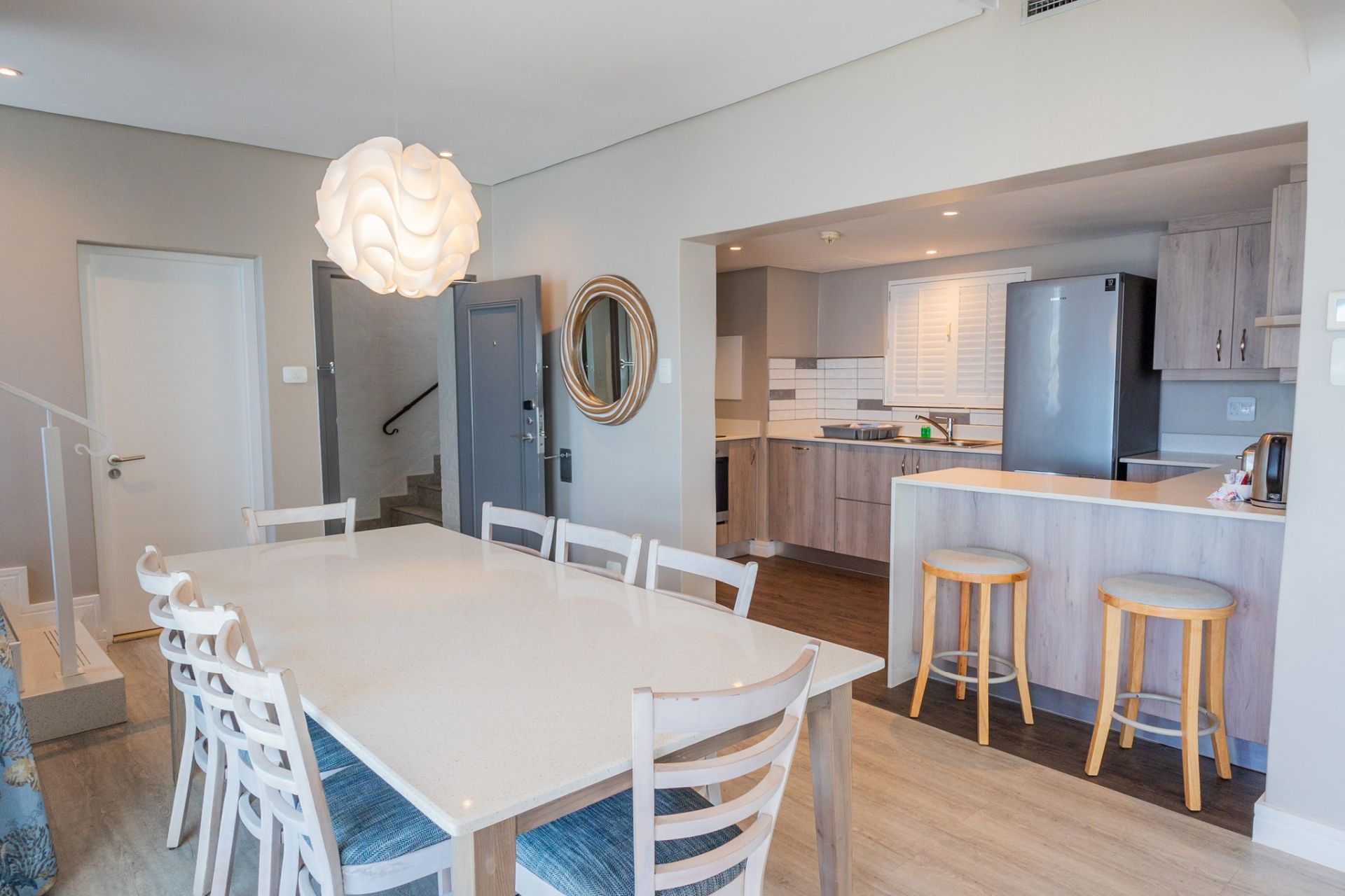 Una cocina moderna con una isla central, un comedor con mesa y sillas blancas, y una escalera que lleva a otro nivel.