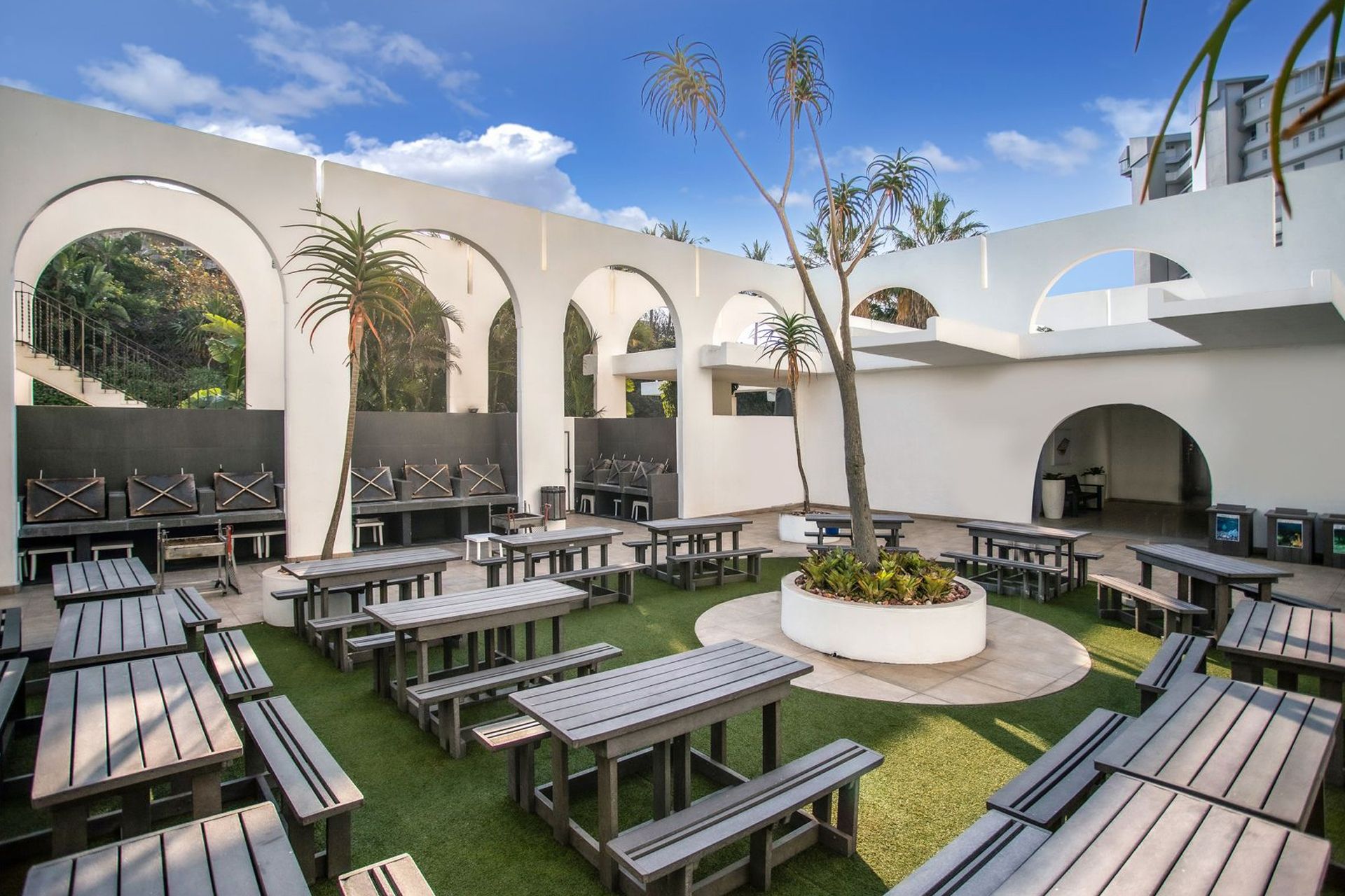 An outdoor patio area with multiple wooden tables and benches, surrounded by white walls with arches, and a central planter with greenery.