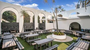 An outdoor patio area with multiple wooden tables and benches, surrounded by white walls with arches, and a central planter with greenery.