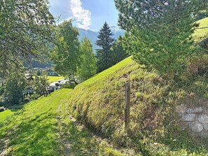 A hillside with a stone wall and a wooden post, surrounded by greenery and houses in the background.