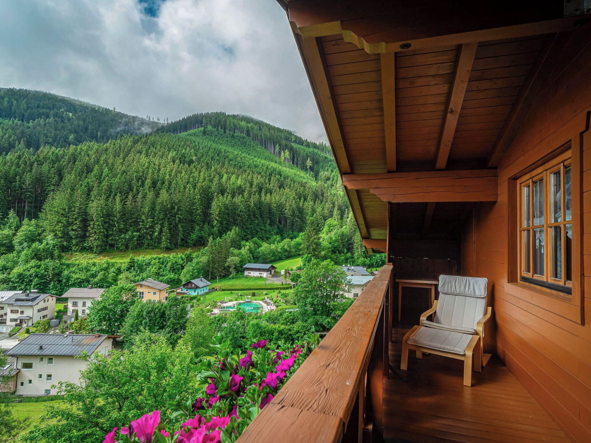 A wooden balcony with a view of a forested valley and houses.