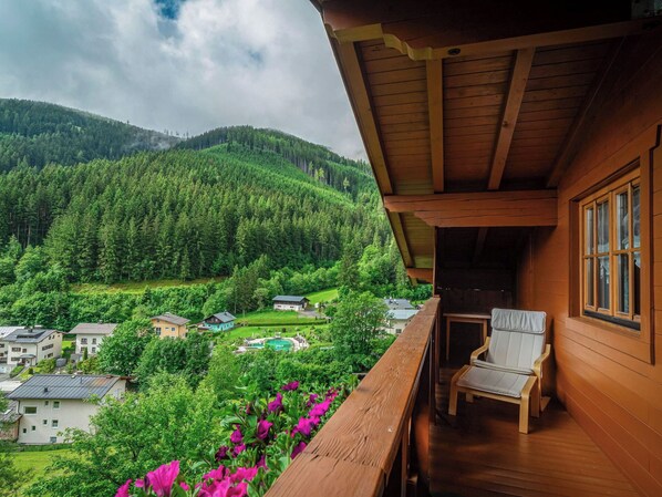A wooden balcony with a view of a forested valley and houses.