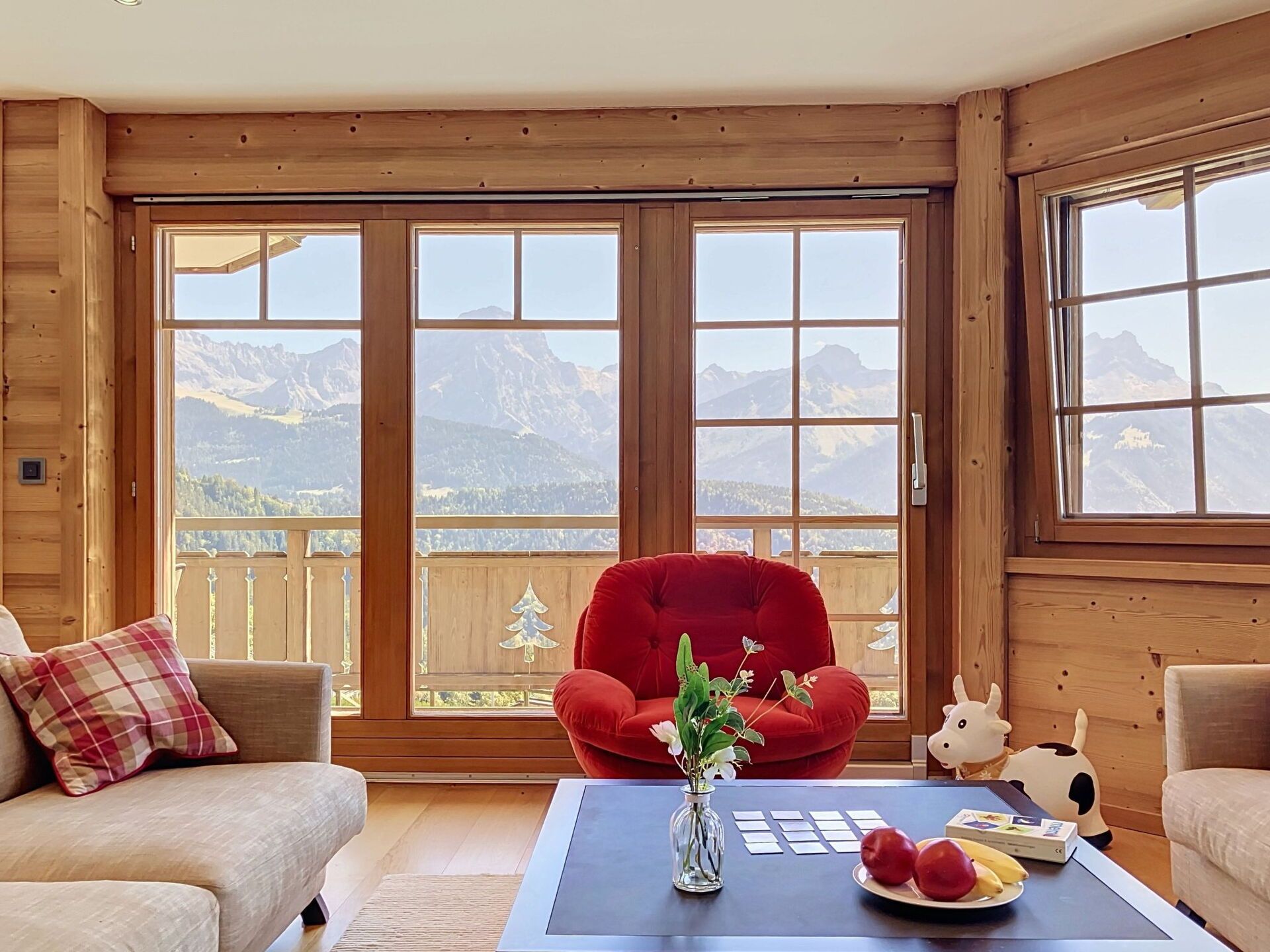 Une pièce chaleureuse avec une grande fenêtre donnant sur les montagnes, un fauteuil rouge, une petite table avec un vase et des fruits, et une figurine de vache.