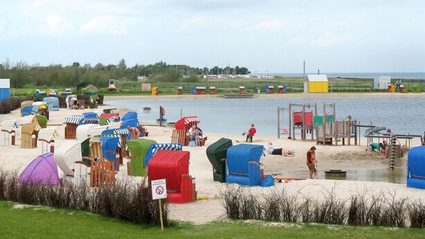 Ein Strand mit vielen bunten Liegestühlen und Sonnenschirmen, einem Spielplatz und ruhigem Wasser im Hintergrund.