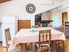 Ein Essbereich mit Tisch, der mit einer blumigen Tischdecke bedeckt ist, Holzstühle, einer Uhr an der Wand, einem weißen Kühlschrank und einem Kamin.