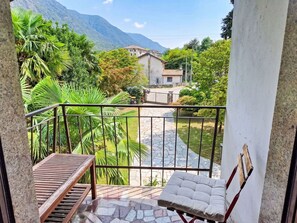 Ein Balkon mit einer hölzernen Bank und einem gepolsterten Stuhl, mit Blick auf einen Garten mit Palmen und einem Gebäude im Hintergrund.