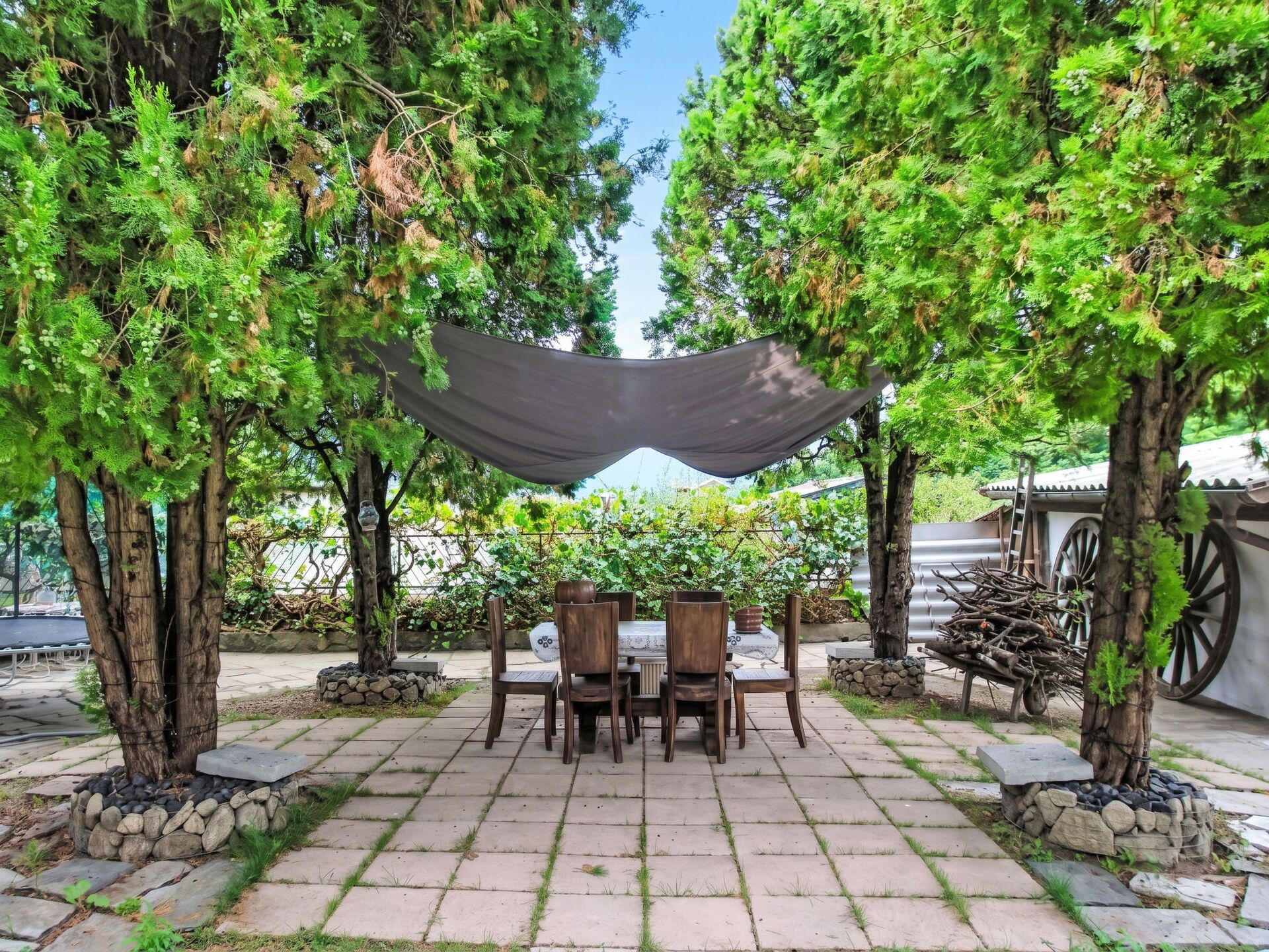 An outdoor patio with a table and chairs, surrounded by trees and a shaded area.